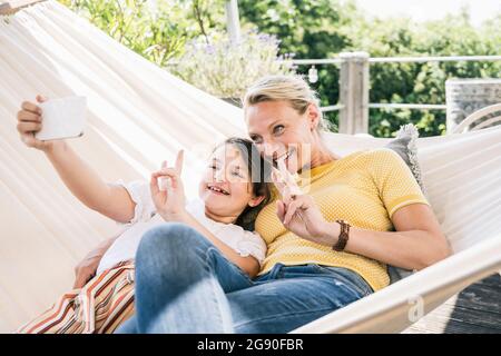 Madre e figlia gesturing segno di pace mentre prendono selfie attraverso il telefono cellulare al balcone Foto Stock