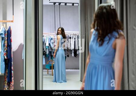 Giovane donna che cerca un abito blu mentre guarda a specchio la boutique Foto Stock