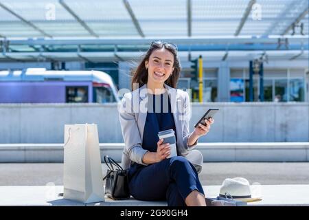 Donna sorridente che tiene la tazza riutilizzabile e lo smartphone mentre si siede sulla parete di ritegno Foto Stock