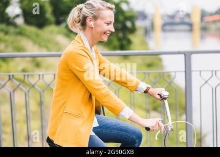 Donna sorridente, con cuffie auricolari wireless che pedalano in bicicletta Foto Stock