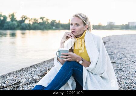 Donna matura pensata che tiene la tazza di caffè mentre si siede vicino al lungofiume Foto Stock