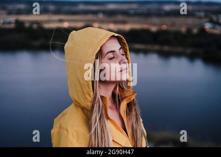 Giovane donna con occhi chiusi indossando un impermeabile giallo al tramonto Foto Stock