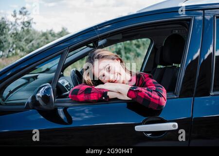 Felice giovane donna appoggiata sulla finestra seduta in auto Foto Stock