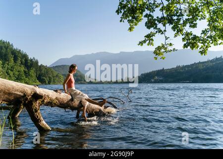 Donna zampe penzolanti e spruzzi d'acqua mentre si siede su albero caduto a Lakeshore Foto Stock