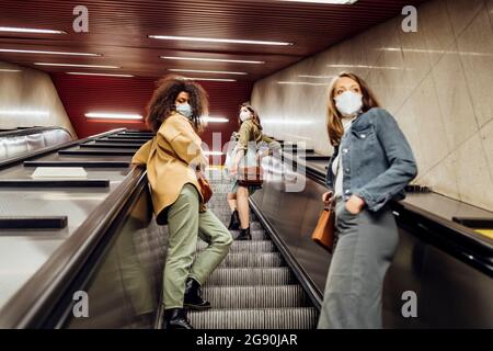 Amici con maschera protettiva in piedi a distanza sociale su scala mobile Foto Stock