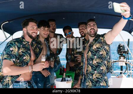 Uomo che attacca la lingua mentre prende selfie con gli amici maschi durante la festa in yacht Foto Stock