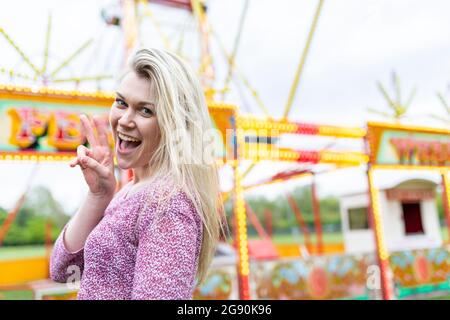 Allegra donna bionda gesturing segno di pace mentre si trova al parco divertimenti Foto Stock