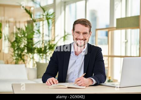 Felice maschio professionale scrittura in diario a tavola mentre lavora da casa Foto Stock