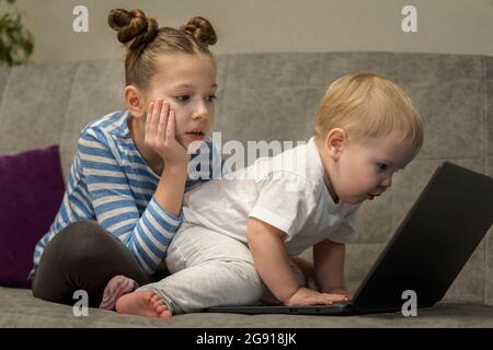 Bambina e ragazzo carino che usano il computer portatile insieme, guardando lo schermo, guardando i cartoni animati o giocando online, sorella e fratello, fratelli seduti su comfor Foto Stock