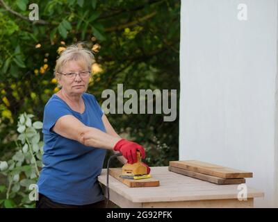 Donna caucasica anziana in una t-shirt blu e guanti rossi sta levigando una tavola con una levigatrice su un layout in un giardino vicino a una parete bianca a casa Foto Stock