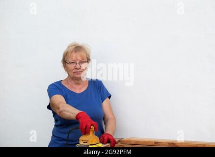 Donna caucasica anziana in una t-shirt blu e guanti rossi sta levigando una tavola con una levigatrice su un layout in un giardino vicino a una parete bianca a casa Foto Stock