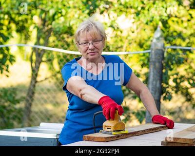 Donna caucasica anziana in una t-shirt blu e guanti rossi sta levigando una tavola con una levigatrice su un layout in un giardino Foto Stock