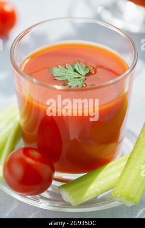 Un bicchiere con succo di pomodoro fresco, sedano, prezzemolo e pomodori maturi su uno sfondo di legno chiaro Foto Stock