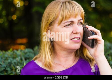 Defocalizzazione primo piano sorridente bionda caucasica che parla, parlando al telefono esterno, all'aperto. donna di 40 anni in blusa viola nel parco. Adulto Foto Stock