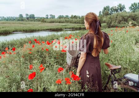 Donna con bicicletta in piedi tra campo papaveri contro il cielo Foto Stock