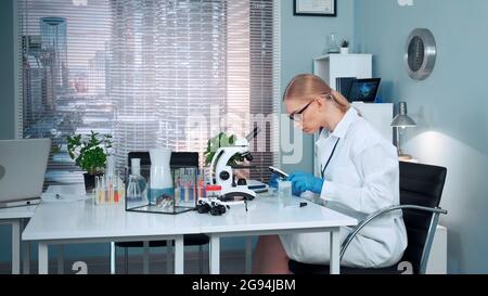 Nel laboratorio di chimica moderna, lo scienziato di ricerca femminile che utilizza la pipetta per far cadere i fertilizzanti sul cursore e poi esaminarlo sotto una lente di ingrandimento Foto Stock