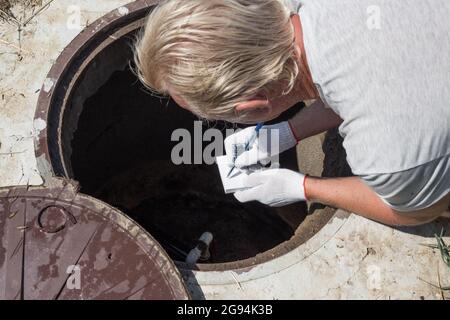 L'uomo si è piegato sopra il pozzo dell'acqua e registra le letture del contatore dell'acqua. Controllo e fissaggio del contatore. Foto Stock