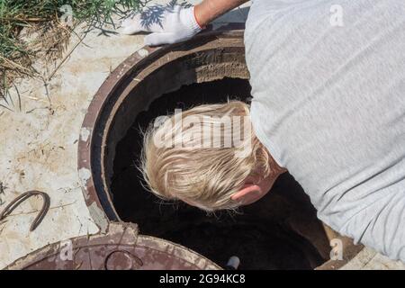 Un uomo si chinò su un pozzo d'acqua e guarda le letture del contatore d'acqua. Controllo e fissaggio del contatore. Foto Stock