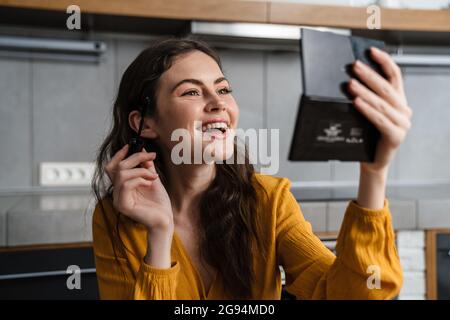 Sorridente giovane donna bruna fare trucco mentre si siede in cucina a casa, tenendo specchio e mascara Foto Stock