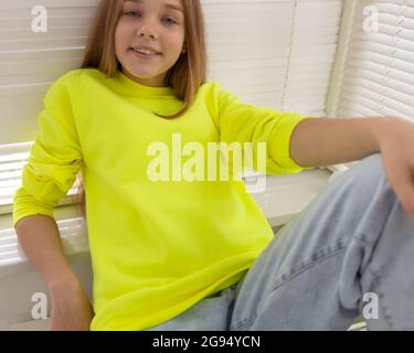 La giovane ragazza adolescente indossa una felpa gialla e si siede davanti alla fotocamera. Sulla sua blusa è spazio di copia per il disegno, l'iscrizione o il logo. Abbigliamento casual per mockup di moda. Foto Stock