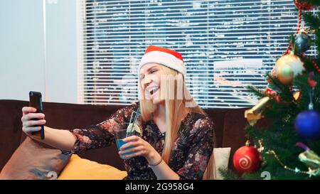 Vista laterale di una donna di buona qualità che fa selfie a Santa Hat e con una bevanda nelle sue mani. Sullo sfondo vi sono un albero di Natale e grattacieli. Foto Stock