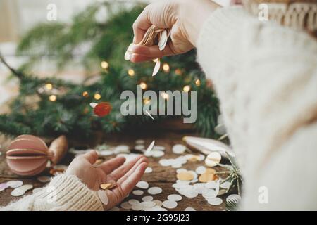 Mani che gettano confetti dorati sullo sfondo di luci natalizie, decorazioni, baubles e rami di pino su legno rustico. Spazio per il testo. Buone vacanze Foto Stock