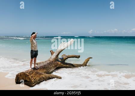 L'uomo si alza sull'albero caduto mentre guarda l'oceano su una spiaggia su Oahu Foto Stock