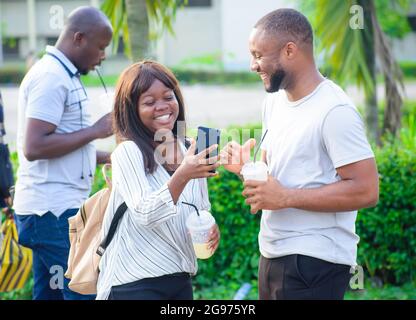 Un gruppo di amici africani composto da due ragazzi e una donna che chiacchiera con gioia con il telefono, parla e godendo il loro tempo libero in un parco all'aperto Foto Stock