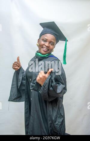 Foto di borsa di una donna africana laureata e/o laureata studenti nel suo vestito nero di laurea e gioiosamente facendo i pollici verso l'alto gesto Foto Stock