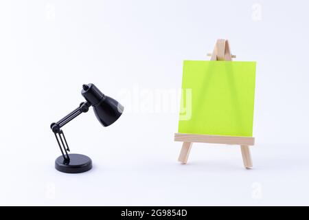 Easel in legno in miniatura con tela quadrata colorata vuota o carta Memo - Mockup. Mini supporto in legno con tavola da disegno pulita e piccola lampada da tavolo nera su sfondo bianco, Copy Space Foto Stock