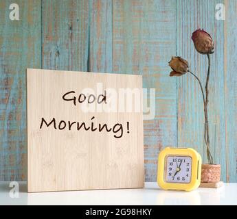 Tavola di legno con il testo Buongiorno con orologio e fiori secchi in vaso sul tavolo. Sfondo di tessitura di legno blu. Foto Stock