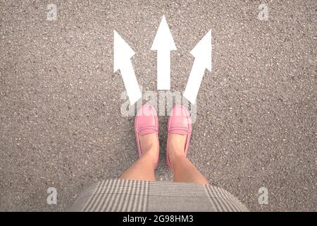 Gambe donna in scarpe rosa in piedi su strada asfaltata con tre frecce bianche di direzione, dritte, a sinistra o a destra. Foto Stock