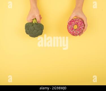 Confronto delle abitudini alimentari. Donna che tiene broccoli e ciambella su sfondo giallo. Spazio di cimasa. Foto Stock