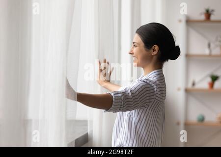 Sorridente sognante donna indiana che apre tende al mattino presto Foto Stock