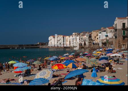 Corpi da spiaggia e luminosi ombrelloni – strisce sedate che si mescolano con fiori, palme e un tocco di tartan – raggruppati sulla sabbia di Cefalù, Sicilia, Italia, sullo sfondo delle pareti bianche e ocra di alte case tegole rosse costruite su resti ad arco delle mura medievali della città. Oltre la baia della città vecchia, gli amanti del sole affollano anche la spiaggia sotto Piazza Marina, il vecchio molo del porto e due arcate in macerie restaurate delle fortificazioni seaward. Foto Stock