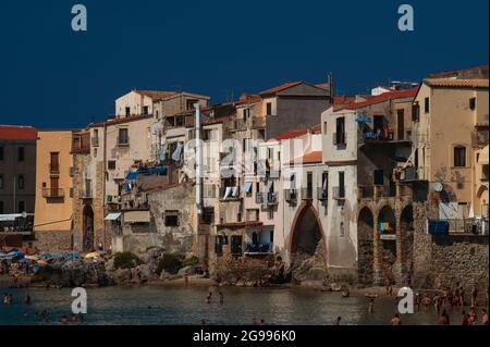 I bagnanti bronzati dal sole si trovano a pale in arrache del Mar Tirreno sotto le mura ocre e bianche delle case del centro storico di Cefalù, Sicilia, Italia, costruite sulle macerie ad arco resti delle mura medievali della città, progettate per proteggere la città costiera settentrionale da attacchi di mare e incursioni piratesche. Cefalù, fondata dagli antichi Greci e occupata a sua volta da Romani, Bizantini, Arabi e Normanni, è diventata una delle località turistiche più famose della Sicilia. Foto Stock