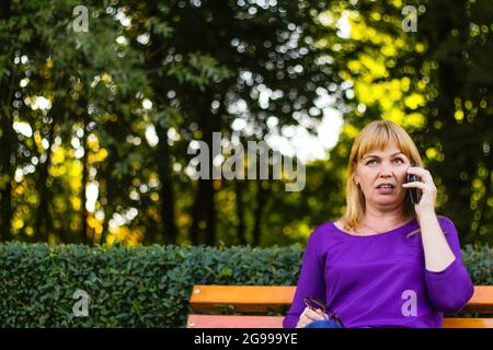 Defocus meraviglia caucasica bionda donna che parla, parlando al telefono esterno, all'aperto. donna di 40 anni in blusa viola nel parco. Persona adulta che utilizza Foto Stock