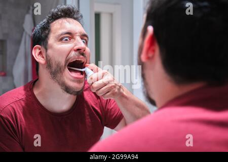 Uomo nel dolore mentre usa uno spray per trattare un'afte o una ferita alla gola davanti allo specchio. Foto Stock