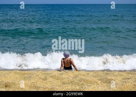 Vacanza, vacanza in spiaggia, estate relax: Donna in costume da bagno nero e cappello luminoso è seduta sulla riva del mare in uno spruzzo di onde di schiuma bianca. Foto Stock