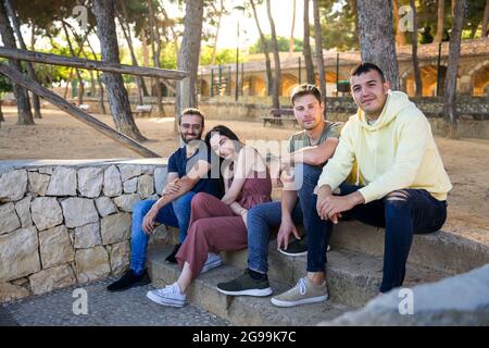 Amici felici seduti sui gradini di un parco con pini. Stanno guardando la macchina fotografica sorridendo. Caucasico, giovani in abiti estivi. Javea Foto Stock