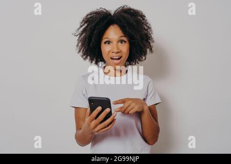 Entusiasta ragazza scura skinned con capelli ricci che tengono il telefono cellulare Foto Stock