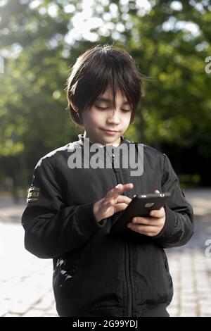 Bimbo ispanico in un parco che gioca con il suo cellulare. Ragazzo ispanico che tiene il cellulare nel parco Foto Stock