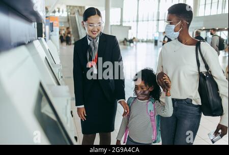 Assistente della compagnia aerea che assiste la ragazza e la madre con macchina self-service per il check-in. Famiglia africana che indossa la maschera all'aeroporto con il personale delle compagnie aeree. Foto Stock