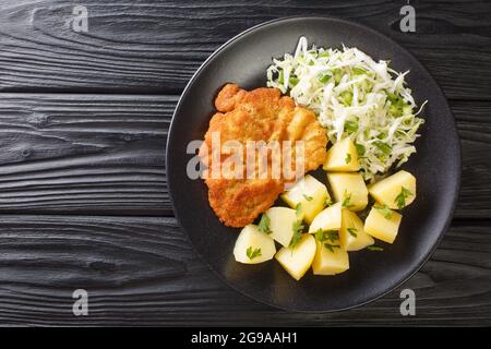 Kotlet Schabowy Polacco maiale cotto con patate bollite e insalata di cavolo primo piano in un piatto su un tavolo di legno nero. Vista orizzontale dall'alto Foto Stock