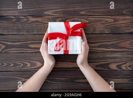 Donna che dà il natale imballato o altro regalo fatto a mano di festa in carta bianca con nastro rosso, tavolo di legno, vista dall'alto Foto Stock