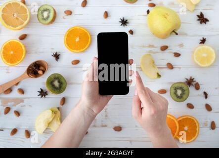 Concetto di dieta. Donna che tiene cellulare con schermo nero vuoto su tavolo di legno con fette di frutta, vista dall'alto, mockup Foto Stock