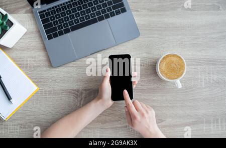 Vista dall'alto dello smartphone femminile che tiene le mani con schermo vuoto, seduto alla scrivania con computer portatile, caffè, blocco note Foto Stock