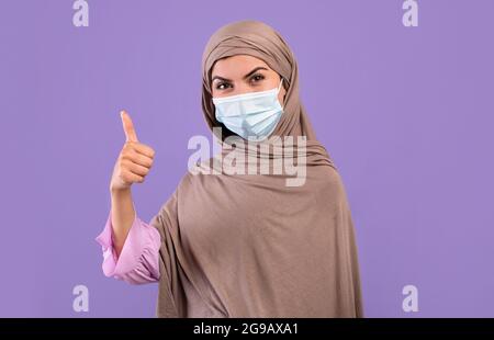 Protezione contro le malattie. Donna armena in hijab indossando una maschera medica protettiva per il viso, mostrando il pollice in su Foto Stock