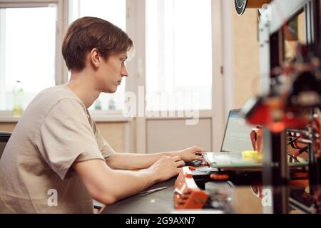 Vista laterale di un giovane maschio seduto al tavolo e creazione di schemi per stampanti 3D su netbook durante il lavoro a casa Foto Stock