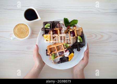 Piatto da donna con waffle belgi decorati con frutta e sciroppo, seduto al tavolo con una tazza di caffè, vista dall'alto Foto Stock